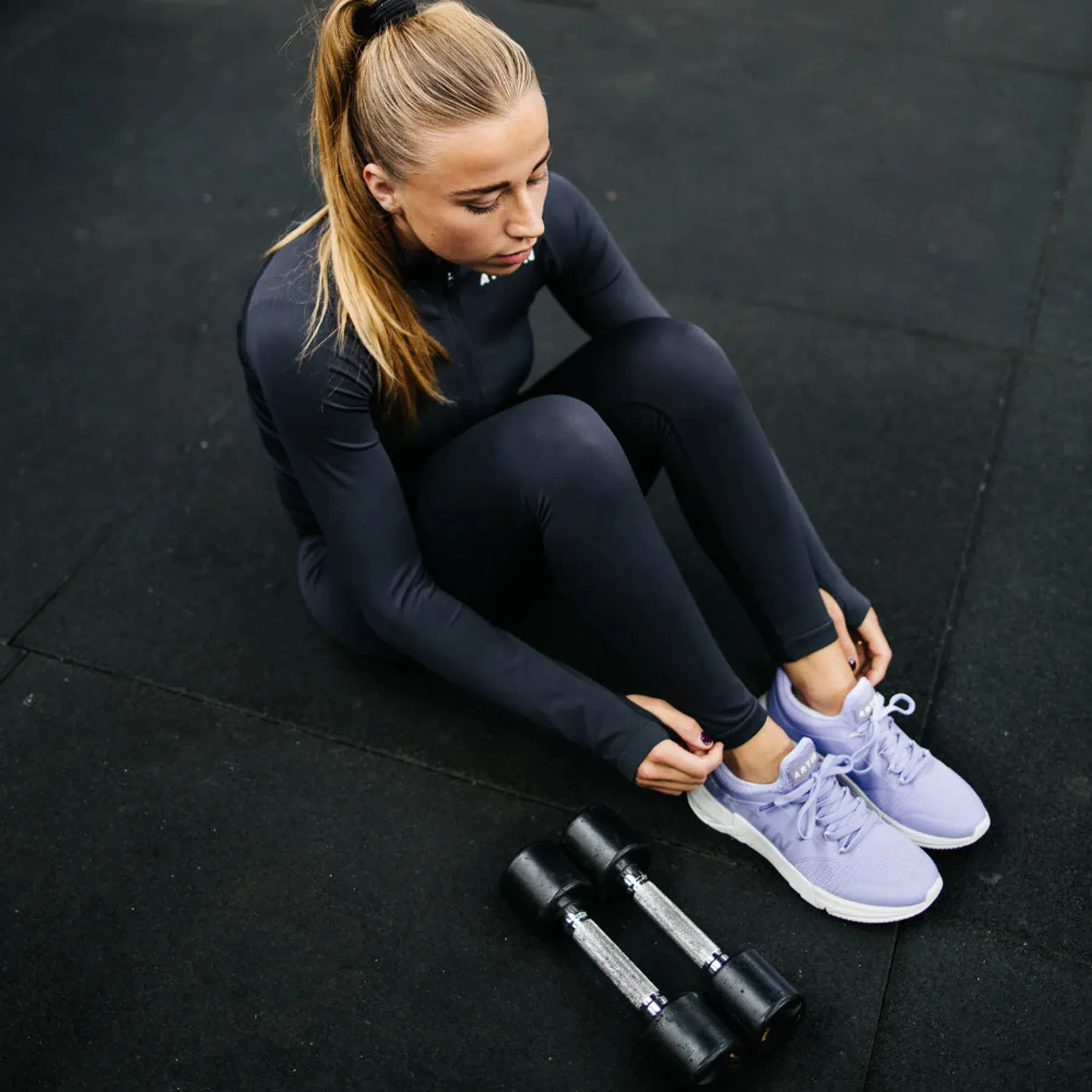 Een vrouw zit op de grond in de sportschool en raakt haar Artin sportschoenen in het paars aan, terwijl naast haar twee gewichten liggen