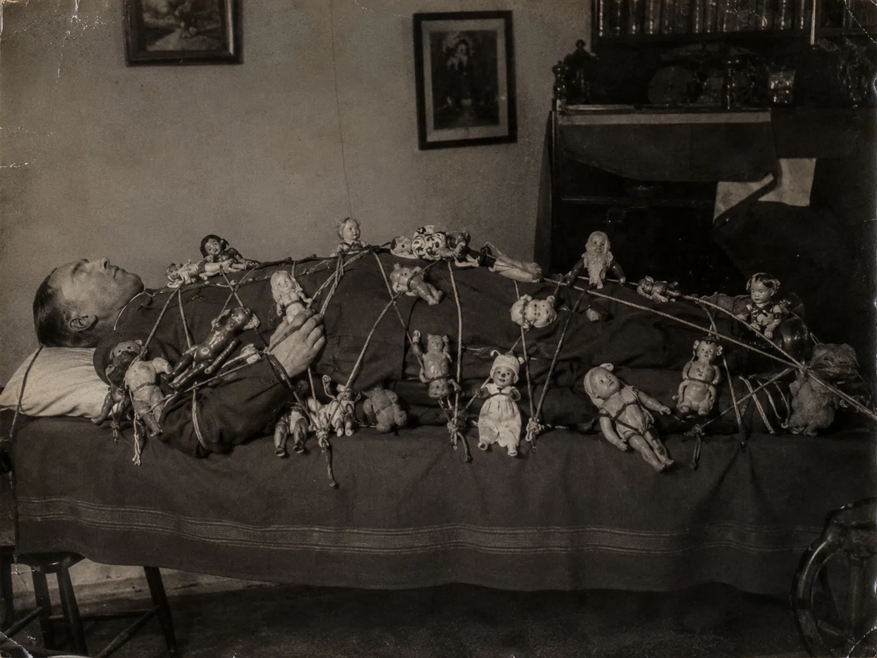 Een (dode?) man op een bed. De man heeft uiteraard geen ogen. Hij is met dunne touwtjes vastgebonden en er liggen tientallen poppen op zijn lichaam. Een daarvan kijkt recht naar de camera en lijkt een beetje op een clown.